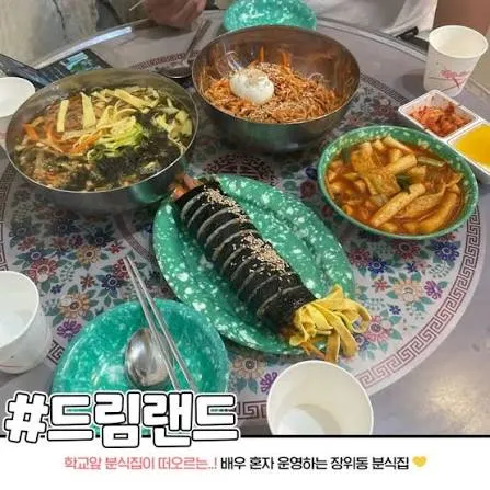 A table spread of Korean bunsik dishes at Dreamland, including a sliced gimbap-style roll, with a Korean social-media caption overlay.