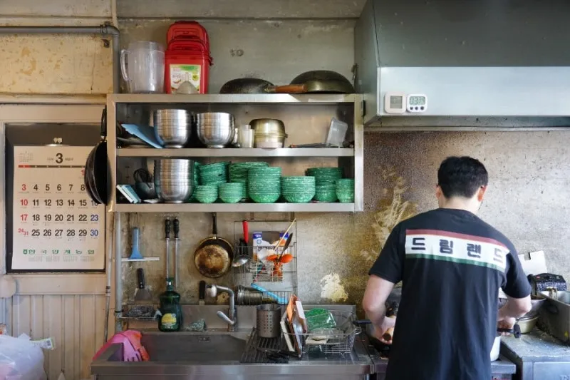 A man with his back to the camera, wearing a t-shirt printed with the restaurant's name, working at a kitchen sink.